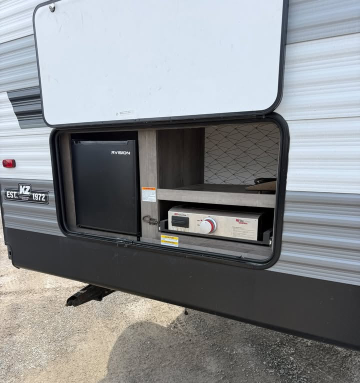 Side view of an RV with a storage compartment open, revealing electronic equipment and fridge.